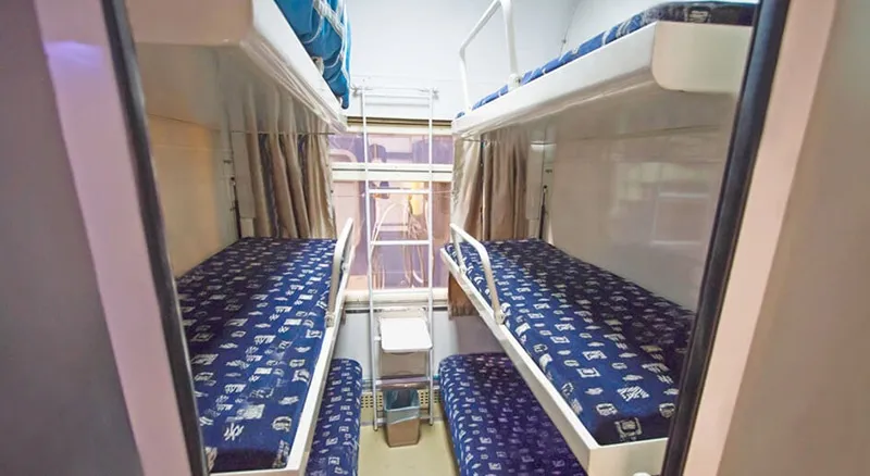 The interior of a six-passenger sleeper train cabin, with three tiers of blue folding berths facing each other across a narrow gangway, a patterned rug on the floor and curtained window at the far end