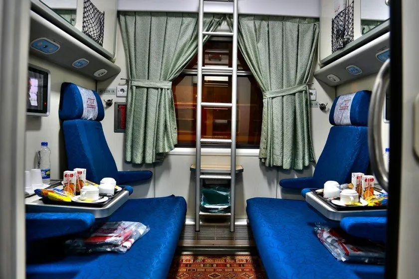 The interior of a four-passenger sleeper train cabin, with two blue facing seats, a folding ladder to the upper berths, green curtains, and trays of refreshments laid out