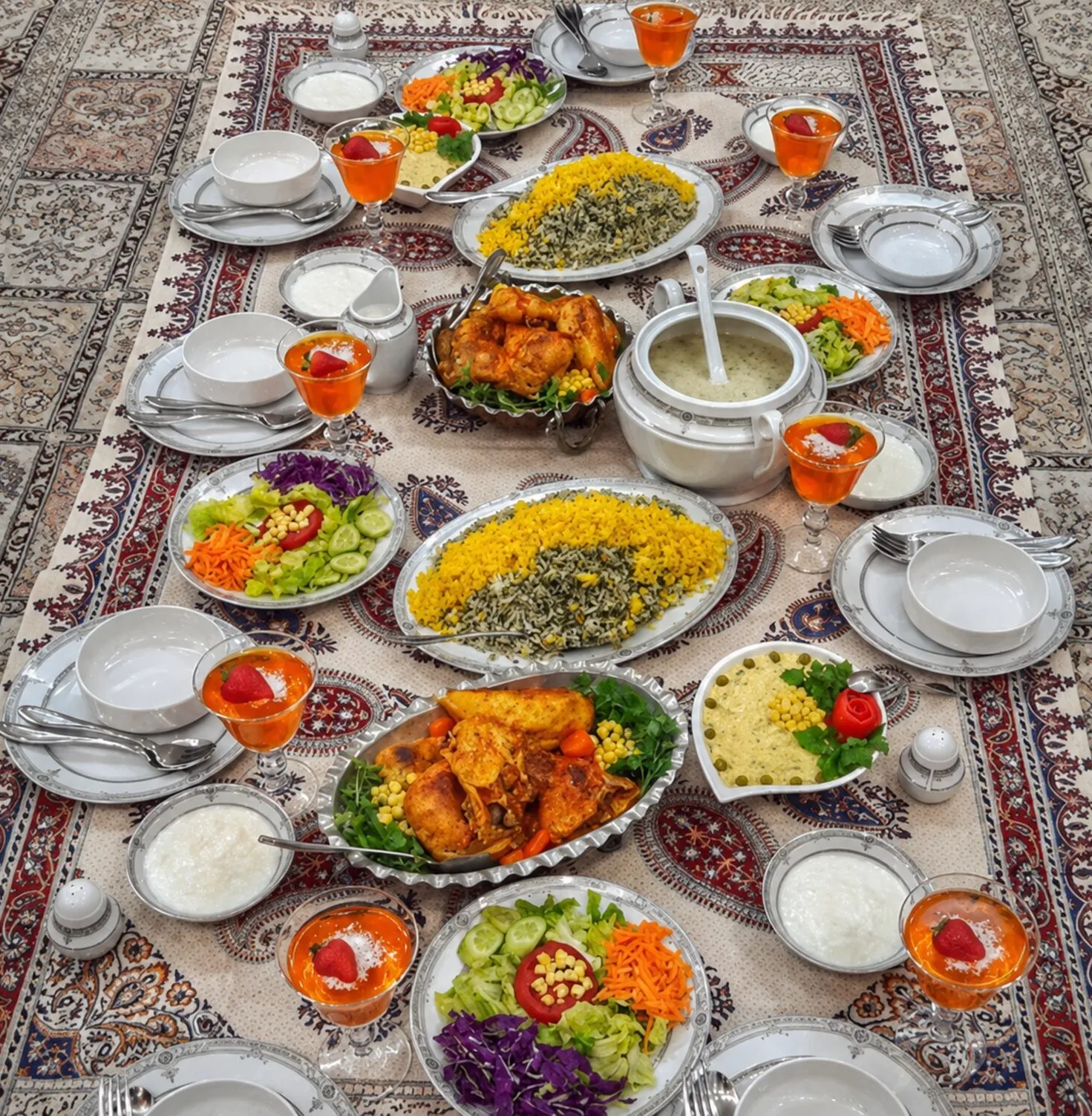 A long sofreh laid over a Persian carpet for a large gathering, set with platters of saffron and herbed rice, roast chicken, stews, bowls of yogurt, salads and glasses of sharbat at every place