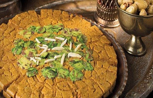 A round Sohan — golden saffron brittle toffee topped with slivered almonds and chopped pistachios — on a copper tray
