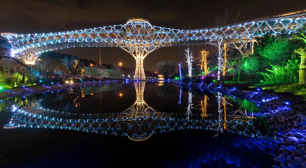The Tabiat (Nature) Bridge in Tehran illuminated at night, a curving three-level steel pedestrian walkway on tree-shaped supports, reflected in still water below