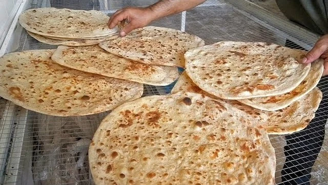A baker lifting a round, thin Taftoon flatbread from a stack of freshly baked breads on a wire cooling rack