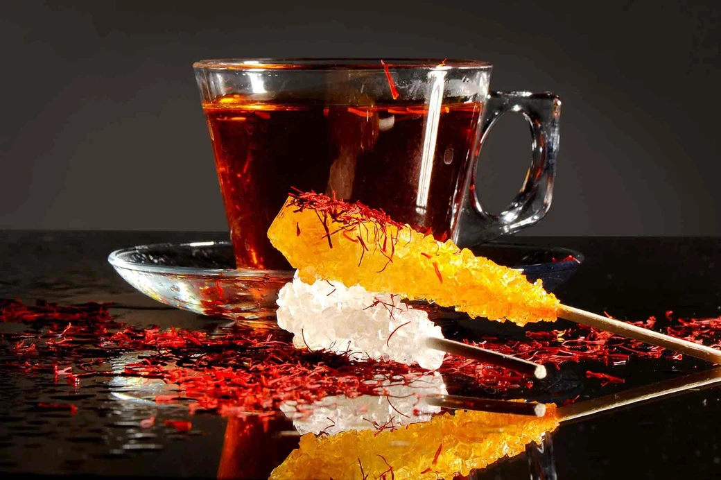 A glass cup of dark Iranian black tea on a saucer, beside sticks of saffron rock candy and scattered saffron threads