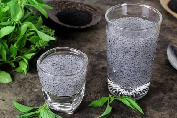 Two glasses of Tokhm Sharbati — translucent basil seeds suspended in water — beside fresh basil leaves and a dish of dry seeds