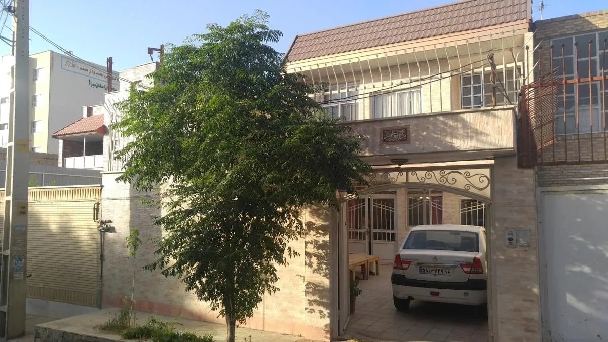A typical two-storey villa-style house on a residential street in Qum, with a brick façade, a balcony, a tree out front and a car parked in the gated carport