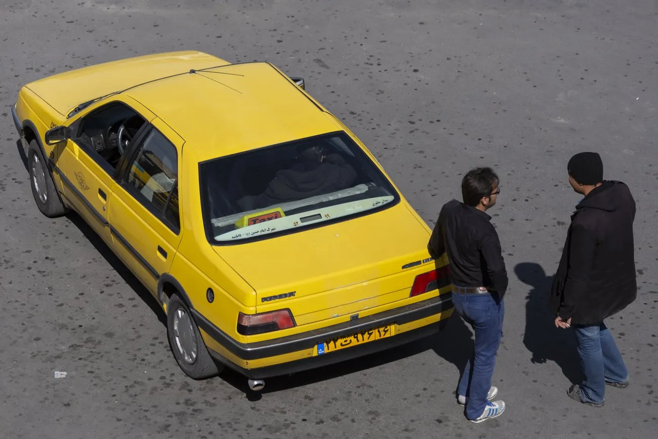 A yellow Peugeot taxi parked on a paved square in Qum, with two men standing beside it negotiating the fare